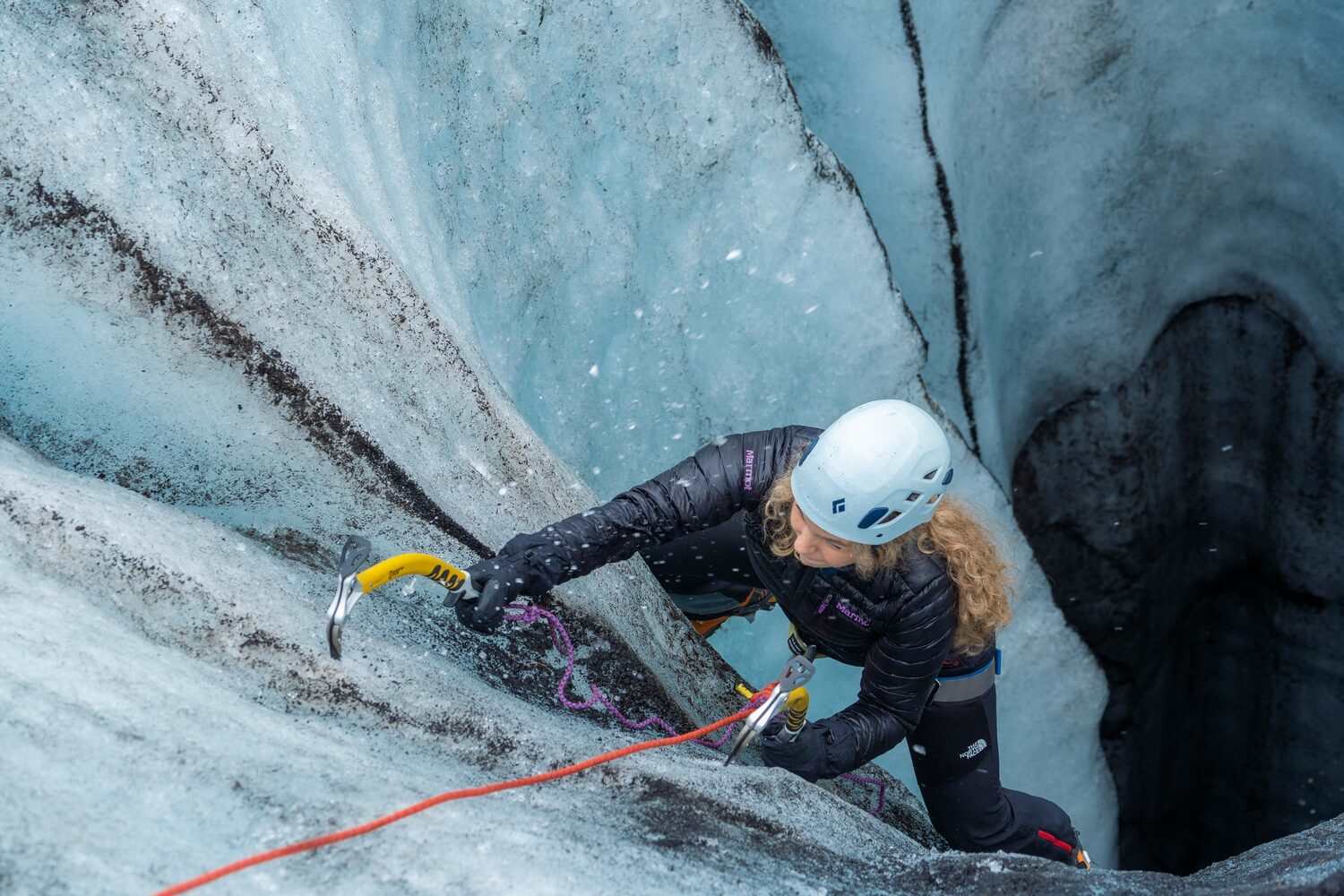 Female in white helmet climbing upwards from ice tunnel with yellow ice axes