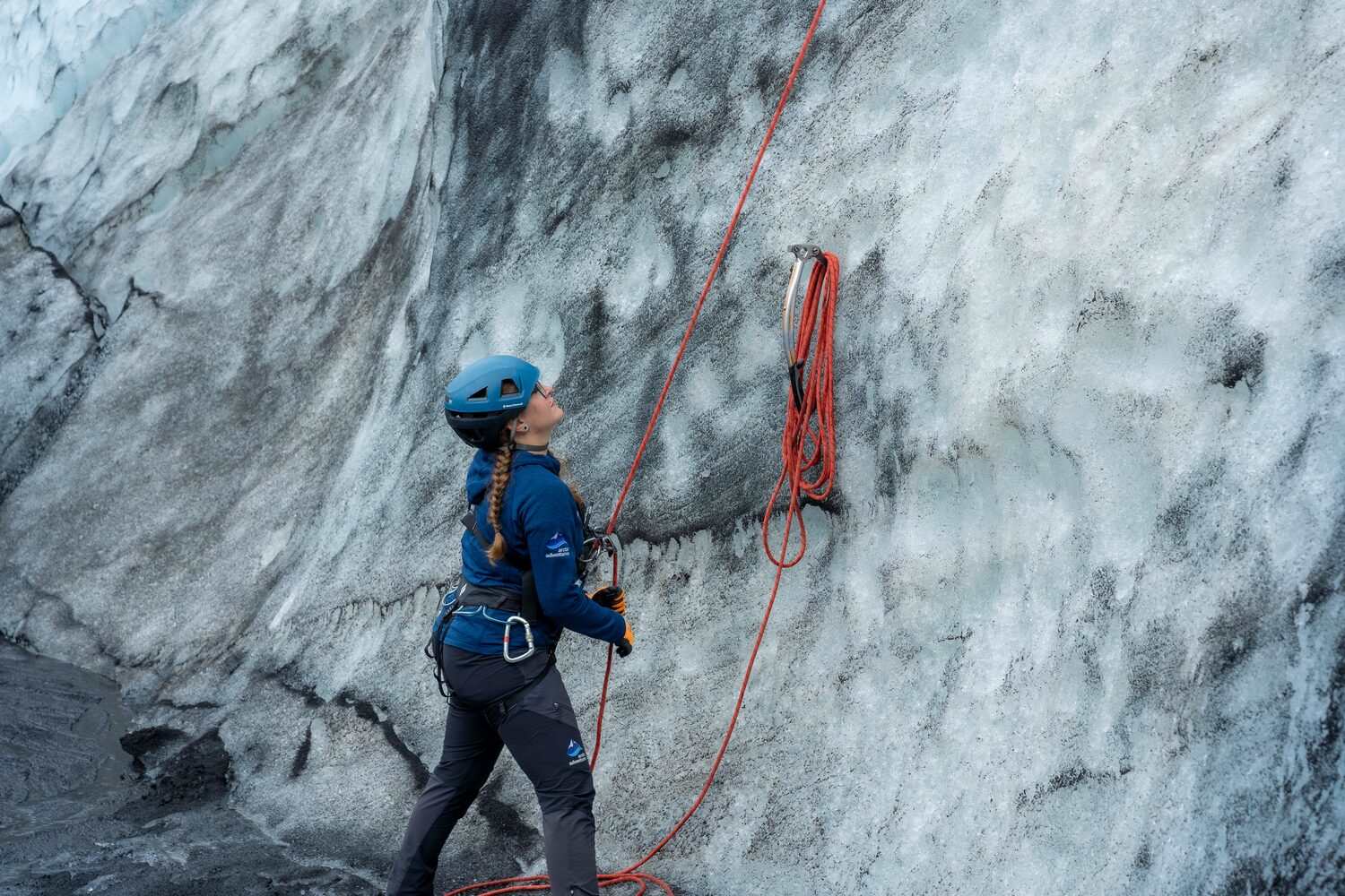 Female with orange safety rope standing at bottom of grey and white ice pattern glacier